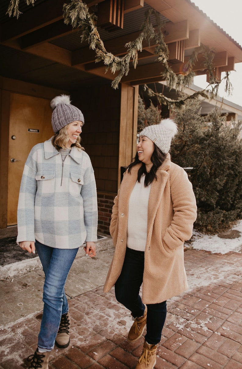May include: Two women wearing winter clothing stand outside a building. One woman is wearing a blue and white plaid shirt, blue jeans, and a gray knit hat. The other woman is wearing a brown teddy bear coat, blue jeans, and a white knit hat. They are both smiling and looking at each other.