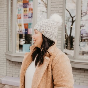 May include: A woman wearing a light brown fuzzy coat with a white knit beanie with a pom pom. The beanie has a textured knit pattern.