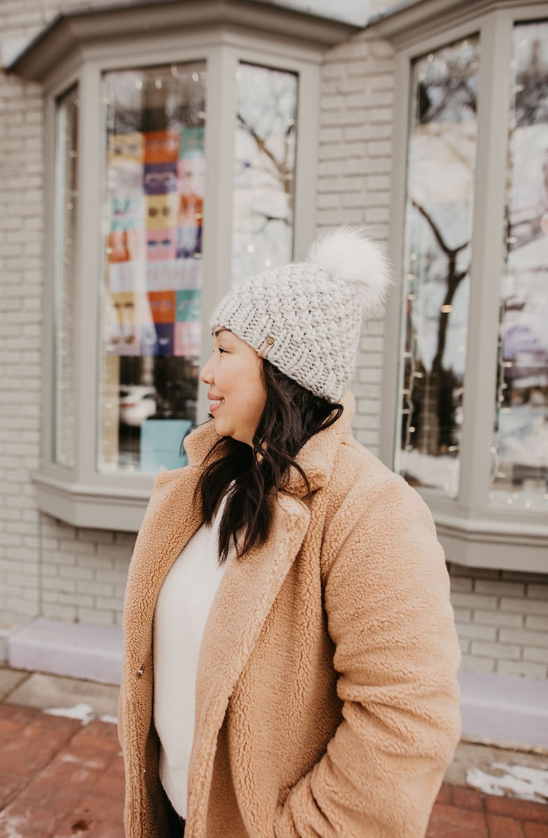 May include: A woman wearing a light brown fuzzy coat with a white knit beanie with a pom pom. The beanie has a textured knit pattern.