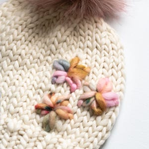 May include: A cream-coloured knitted hat featuring a large, fluffy, mauve pom-pom. The hat is decorated with three colourful, embroidered flower appliques in shades of pink, yellow, and green. The knit has a chunky texture.