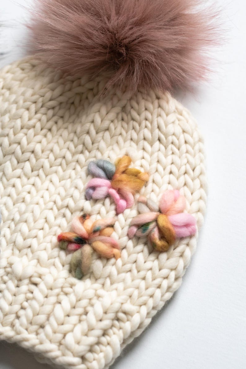 May include: A cream-coloured knitted hat featuring a large, fluffy, mauve pom-pom. The hat is decorated with three colourful, embroidered flower appliques in shades of pink, yellow, and green. The knit has a chunky texture.