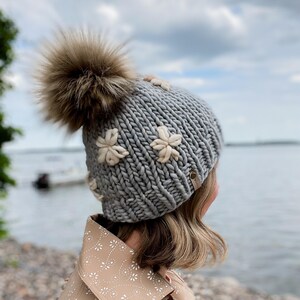 May include: A grey knitted beanie featuring cream flower appliqu&eacute;s and a large brown fur pom-pom. The hat is made of thick yarn and has a small tag on the side. A winter accessory.