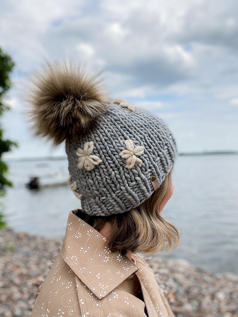 May include: A grey knitted beanie featuring cream flower appliqu&eacute;s and a large brown fur pom-pom. The hat is made of thick yarn and has a small tag on the side. A winter accessory.