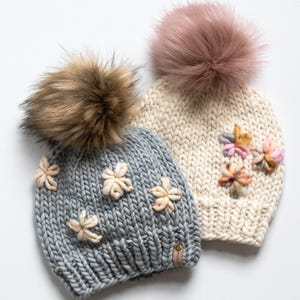 May include: Two knitted beanies with faux fur pom-poms. One is light grey with cream-coloured flower accents, the other is cream-coloured with pink and peach flower accents. Both have a small leather tag.