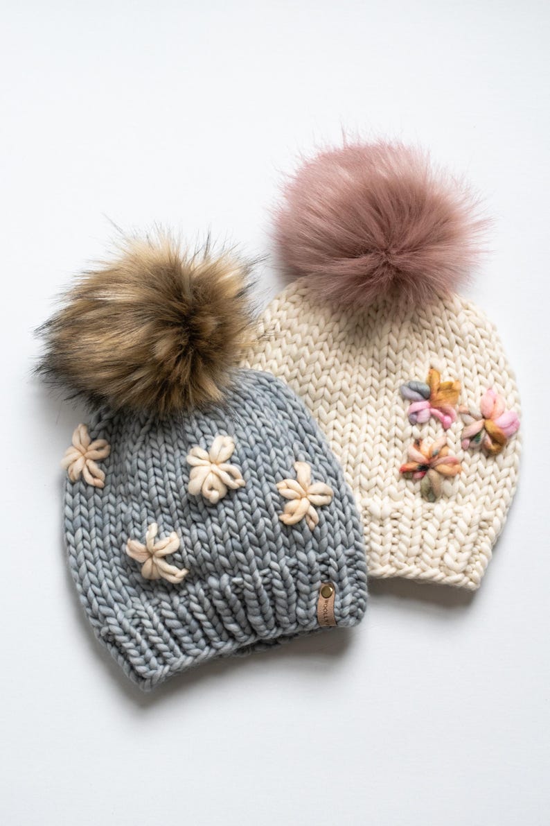 May include: Two knitted beanies with faux fur pom-poms. One is light grey with cream-coloured flower accents, the other is cream-coloured with pink and peach flower accents. Both have a small leather tag.