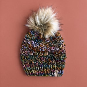 May include: A colorful knitted beanie with a faux fur pom pom. The beanie has a brown leather tag with the word "WOOL" on it.