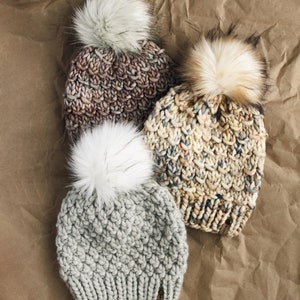 May include: Three knitted winter hats with faux fur pom poms. The hats are in shades of gray, brown, and beige.