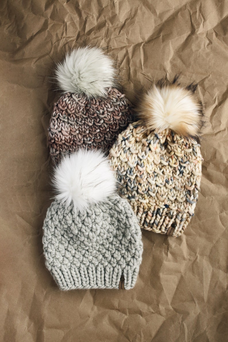 May include: Three knitted winter hats with faux fur pom poms. The hats are in shades of gray, brown, and beige.