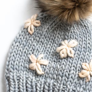 May include: A pale blue knitted hat with a large brown faux fur pom-pom and several cream-coloured flower embellishments. The hat is made of thick, chunky yarn, and the flowers are three-dimensional.