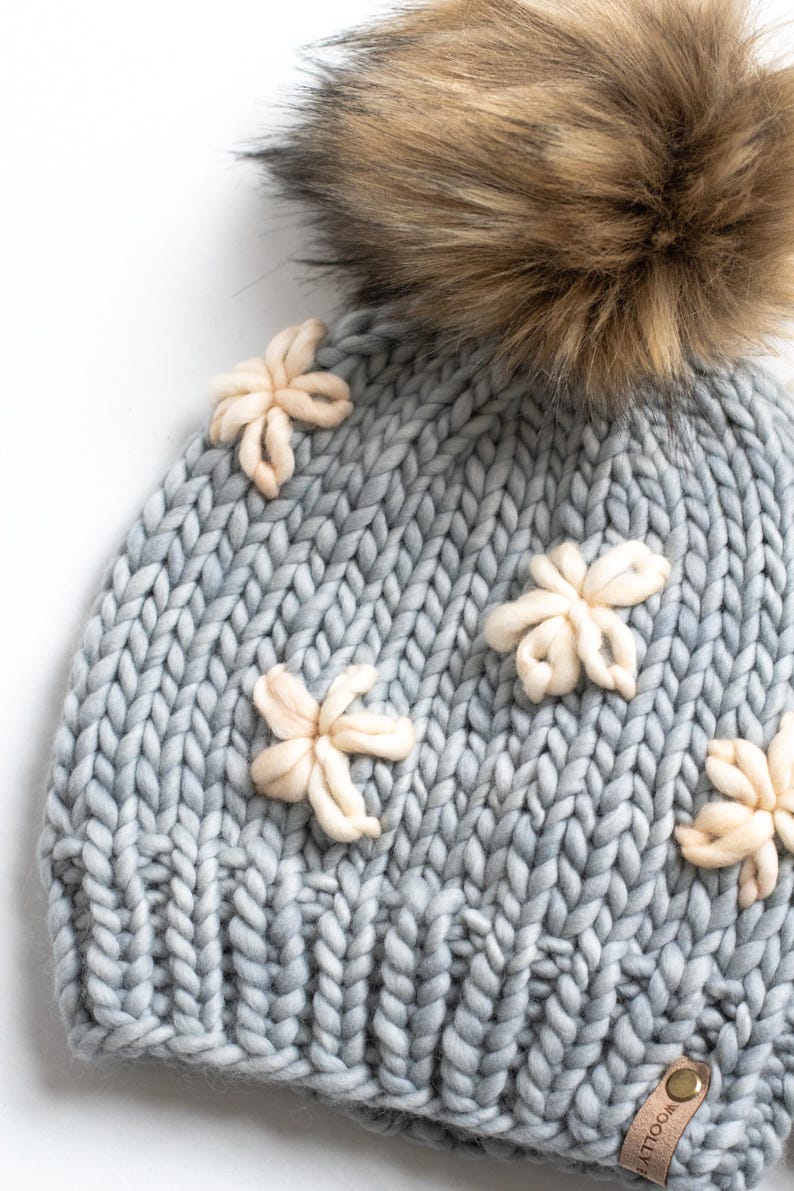 May include: A pale blue knitted hat with a large brown faux fur pom-pom and several cream-coloured flower embellishments. The hat is made of thick, chunky yarn, and the flowers are three-dimensional.