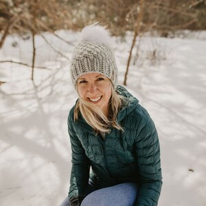 May include: A woman wearing a teal blue puffer jacket, a grey knit beanie with a white pom pom, and blue jeans. She is sitting in the snow with her hands tucked inside her jacket.