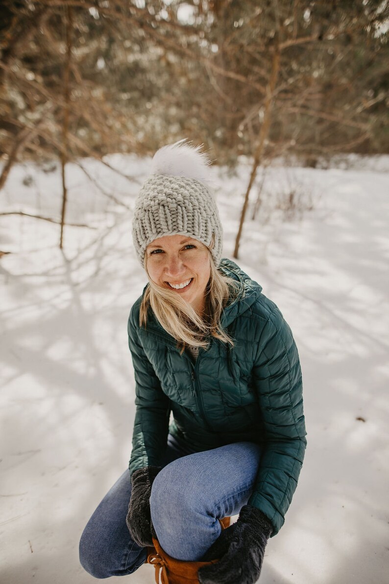 May include: A woman wearing a teal blue puffer jacket, a grey knit beanie with a white pom pom, and blue jeans. She is sitting in the snow with her hands tucked inside her jacket.