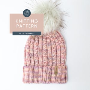 Puede incluir: Un gorro de punto rosa y blanco con un pompón blanco esponjoso. El gorro tiene un patrón de punto de cable y una etiqueta de cuero marrón que dice "Woolly Bear Knits".