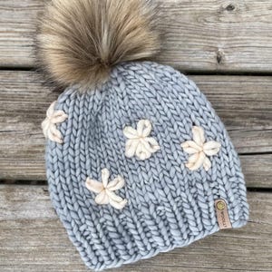 May include: A pale blue knitted beanie featuring cream-coloured flower appliqu&eacute;s and a large, fluffy, brown faux fur pom-pom. The hat has a small, brown leather tag. The beanie is made of thick, chunky yarn.