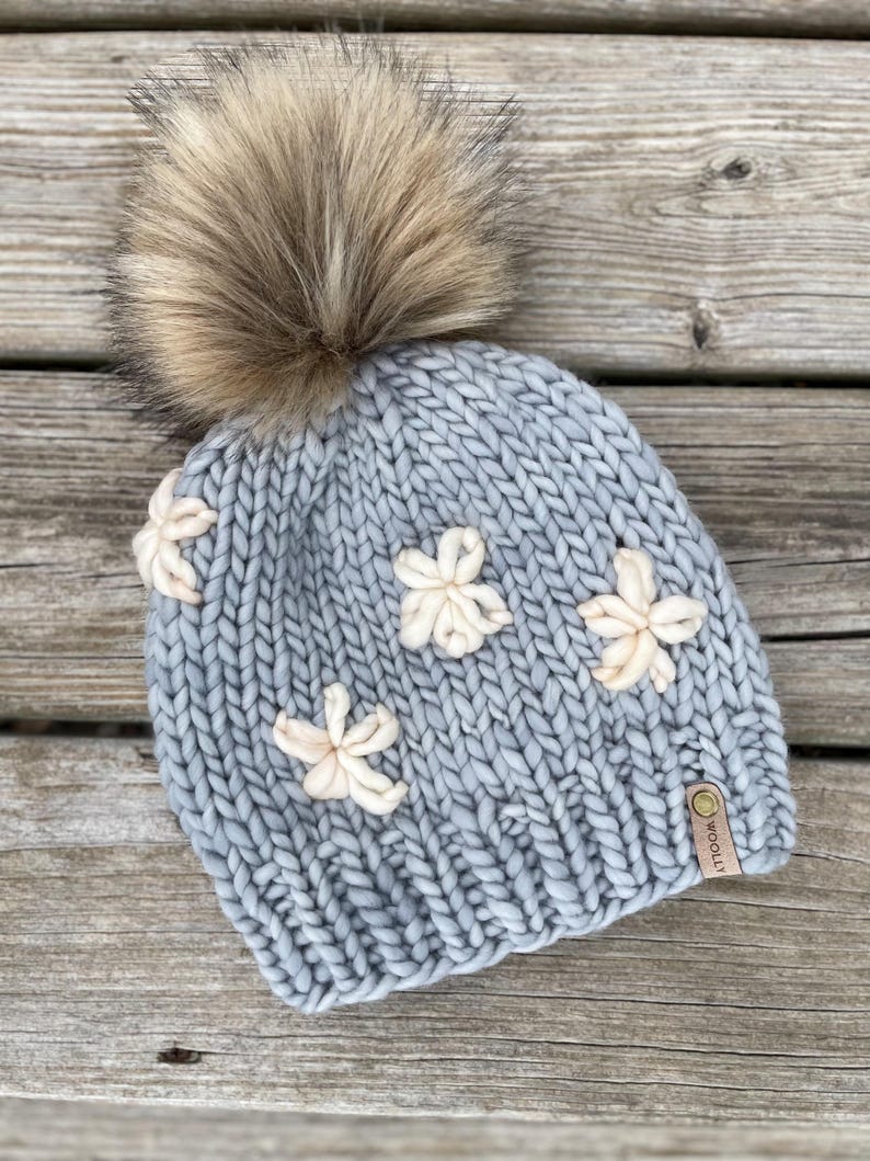 May include: A pale blue knitted beanie featuring cream-coloured flower appliqu&eacute;s and a large, fluffy, brown faux fur pom-pom. The hat has a small, brown leather tag. The beanie is made of thick, chunky yarn.