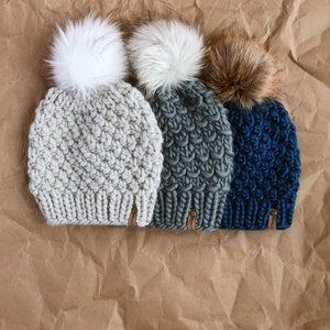May include: Three knitted hats with faux fur pom poms. The hats are in gray, green, and blue. The pom poms are white, white, and brown.