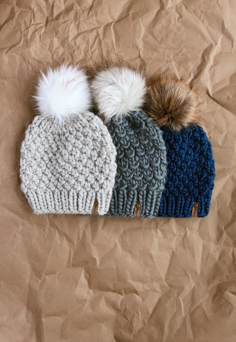 May include: Three knitted hats with faux fur pom poms. The hats are in gray, green, and blue. The pom poms are white, white, and brown.