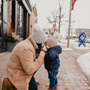 May include: A woman and a child wearing winter hats are standing on a sidewalk in front of a building. The woman is wearing a brown coat and the child is wearing a blue jacket. They are both looking at each other.