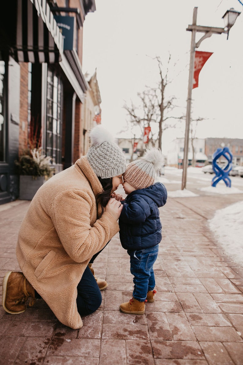 May include: A woman and a child wearing winter hats are standing on a sidewalk in front of a building. The woman is wearing a brown coat and the child is wearing a blue jacket. They are both looking at each other.