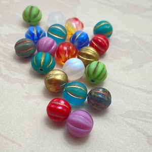 Puede incluir: Una colección de cuentas redondas y coloridas en varios tonos de verde, azul, rojo, dorado y morado. Cada cuenta tiene una textura acanalada y rayas doradas. Las cuentas están dispersas, mostrando sus diversos colores y diseños, ideales para la creación de joyas.