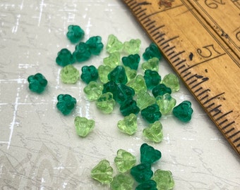 Czech glass blossom flower beads in shades of green and teal scattered on a white surface next to a wooden ruler for scale.