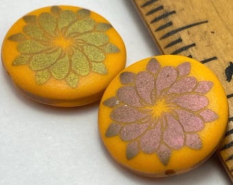 Two matte orange Czech glass coin beads with laser etched flower designs in purple, resting in a dark wooden dish.