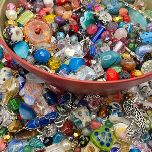 May include: A large collection of assorted loose beads and metal charms in various shapes, sizes, and colours, including glass, stone, and plastic materials, contained within a round red metal tin and scattered on the surface below.