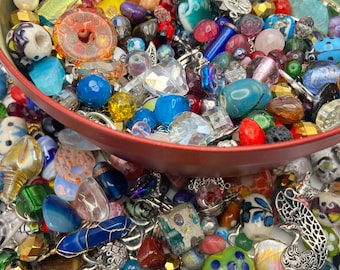 Puede incluir: Una gran colección de cuentas sueltas y dijes de metal variados en diversas formas, tamaños y colores, incluyendo materiales de vidrio, piedra y plástico, contenidos en una lata redonda de metal rojo y esparcidos por la superficie.