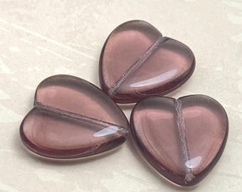 Three amethyst purple Czech glass heart beads on a plain cream background, showing their translucent finish and shiny centre.