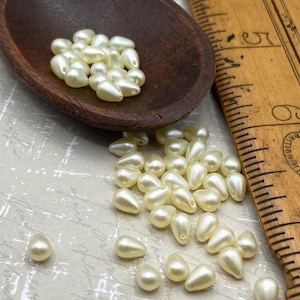 May include: A wooden spoon filled with off-white pearlescent teardrop-shaped beads. The beads are scattered on a beige surface next to a wooden ruler.