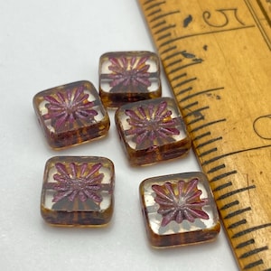May include: Five square glass beads with a clear centre and a dark amber-brown border. Each bead features a deep magenta floral design. A wooden ruler is in the background.