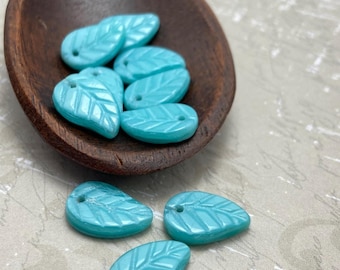 Czech glass leaf beads in turquoise blue, flat leaf-shaped with veined detail, some in a wooden spoon and some on a textured.