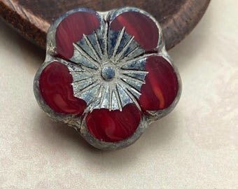 A single deep red Czech glass hibiscus flower bead with antique silver finish lies on a pale background, showing five.
