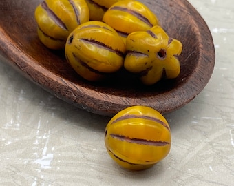 Six pumpkin yellow Czech glass melon beads with brown wash detail, five in a wooden dish and one on white surface.