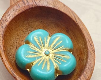 A turquoise sea green Czech glass hibiscus flower bead with gold wash details sits in a wooden spoon against a neutral.