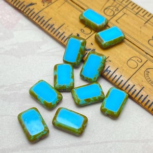 May include: Rectangular beads with a vibrant turquoise centre and a distressed, olive green border. The beads are scattered on a wooden ruler, highlighting their unique colour and design. These beads are suitable for jewellery making.