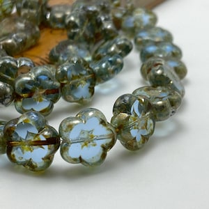 Puede incluir: Hilo de cuentas con forma de flor en tonos azules, verdes y dorados. Cada cuenta presenta un centro azul claro con un diseño floral blanco. Las cuentas están ensartadas, creando un elemento delicado y decorativo.