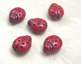 Five red Czech glass ladybird beads with black spots and markings, displayed on a plain cream background.