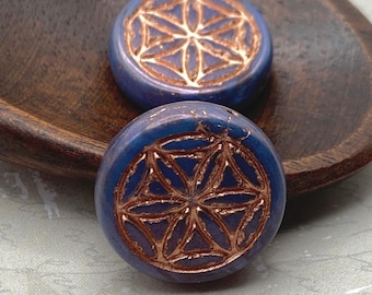 Two violet blue Czech glass Flower of Life beads with metallic copper wash detailing rest on a wooden dish.