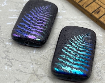 Two matte black Czech glass rectangle beads with holographic fern leaf etchings in purple and cyan, shown beside a wooden.