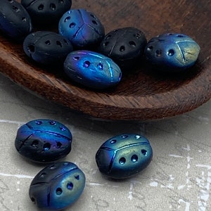 Op de afbeelding: Een verzameling kleine, ovaalvormige kralen die zijn gevormd als kevers. De kralen hebben een matzwarte afwerking of een iriserende metallic blauwe en groene glans, met kleine ingedrukte stippen op de rug om het uiterlijk van insectenvleugels na te bootsen.
