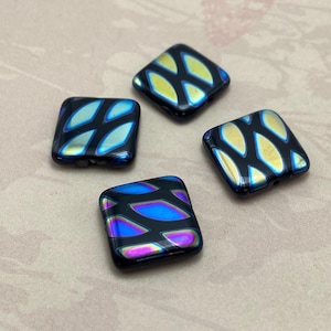 May include: Four square beads with a black base and iridescent, leaf-like patterns in shades of blue, gold, and purple. The beads have a glossy finish and are arranged on a neutral background. These beads are suitable for jewellery making.