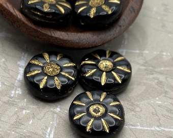Six black Czech glass sunflower beads with gold wash details displayed on a brown wooden dish and linen cloth.