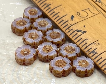 Ten lilac and bronze Czech glass hibiscus flower beads arranged on a white surface next to a wooden ruler for scale.