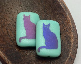 Two matte turquoise laser etched rectangle beads with purple and blue cat silhouettes, displayed in a wooden dish.