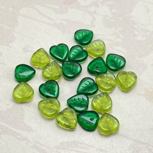 Puede incluir: Una colección de cuentas de vidrio verdes y amarillas en varias formas de hojas y corazones. Las cuentas translúcidas presentan detalles grabados de venas de hojas. Las cuentas están dispersas sobre una superficie clara.