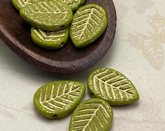 Eight peridot green Czech glass leaf beads with gold wash and etched detail, arranged in a wooden dish and on a plain.