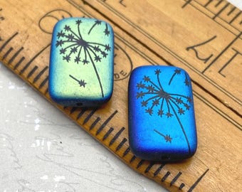 Two laser etched Czech rectangle beads in matte blue with black dandelion design, shown against a wooden ruler for scale.
