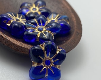 Five sapphire blue Czech glass puffed daisy flower beads with gold detailing, shown in a rustic dish against a light.