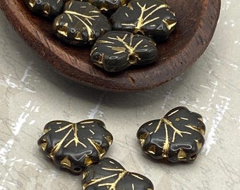 Ten black marble Czech glass maple leaf beads with gold detailing displayed on a wooden dish and plain surface.